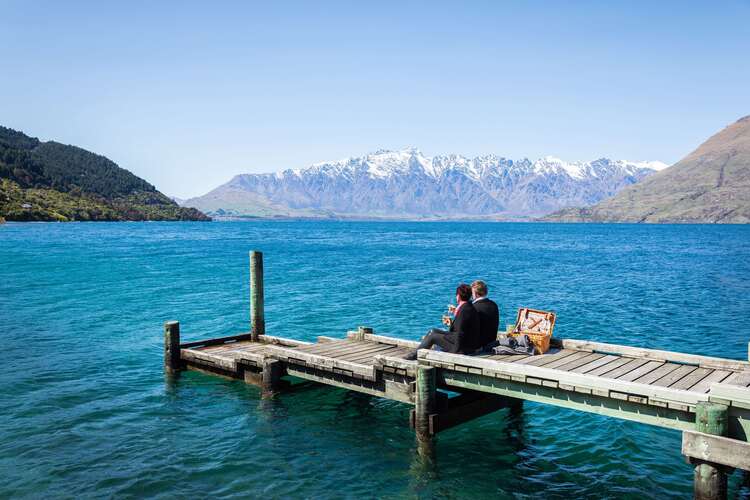Couple sitting on a private dock overlooking a turquoise lake and snow-capped mountains on a luxury girlfriend trip to New Zealand.