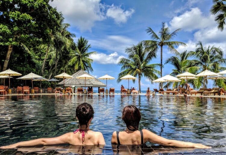 Two women relaxing in a luxury infinity pool on a girlfriend trip, with palm trees and turquoise ocean in the background.