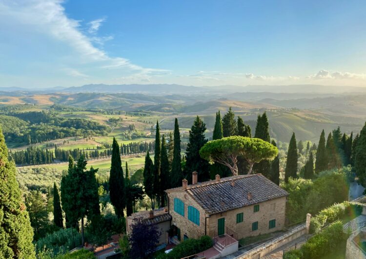 Scenic view of Tuscany’s rolling hills, cypress trees, and a traditional villa under a clear blue sky at sunset.