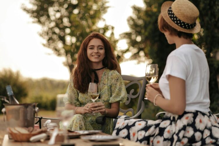 Two women enjoying wine on a girlfriend trip in North America, surrounded by lush greenery in a garden terrace setting.
