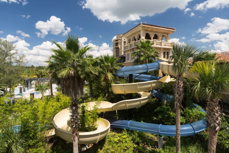 Curving water slides surrounded by palm trees at a resort with Mediterranean-style architecture in the background