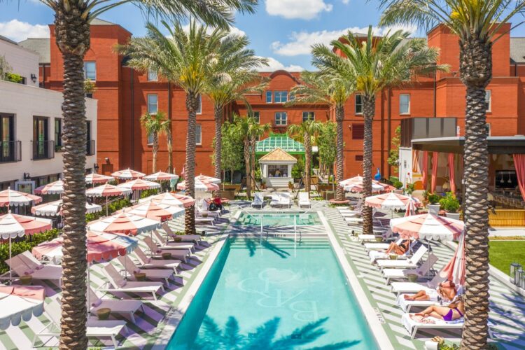 Poolside view at Hotel Bardo Savannah featuring striped loungers, pink umbrellas, palm trees, and red-brick architecture under a blue sky.