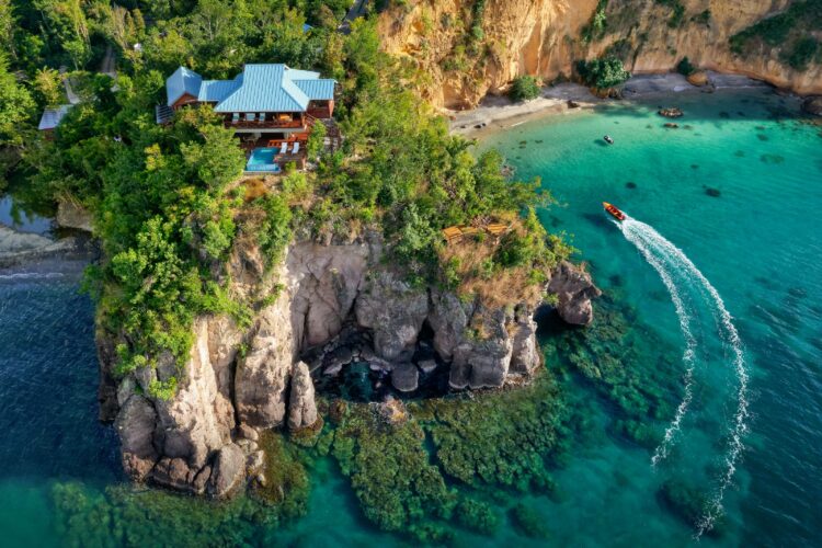 Aerial view of Secret Bay, Dominica, showing a cliffside luxury villa and private plunge pool above a secluded turquoise Secret Beach with a boat cutting through the clear water.